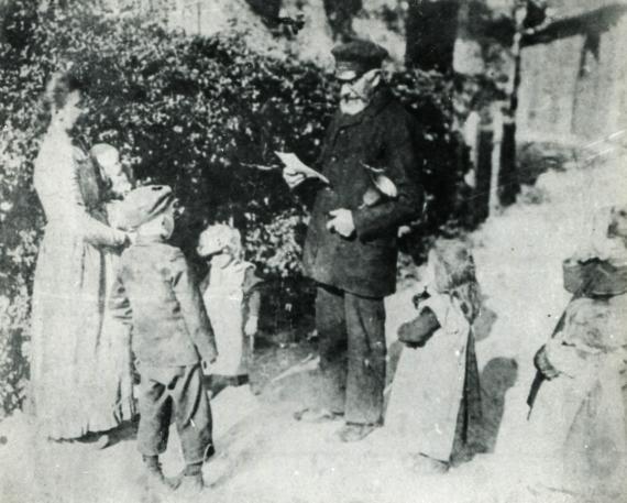 Schwarz-Weiß Fotografie von Heinrich Sauerland, Amtsdiener der Stadt Telgte. Er steht in der Mitte und verliest einen Brief. Um ihn herum steht eine Frau mit Kindern. 