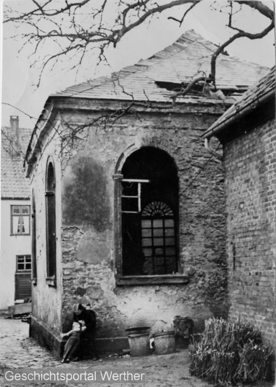 Rückansicht der ehemaligen Synagoge mit Schäden der Putzfassade und Dachdeckung sowie fehlendem Fenster, davor zwei Kinder