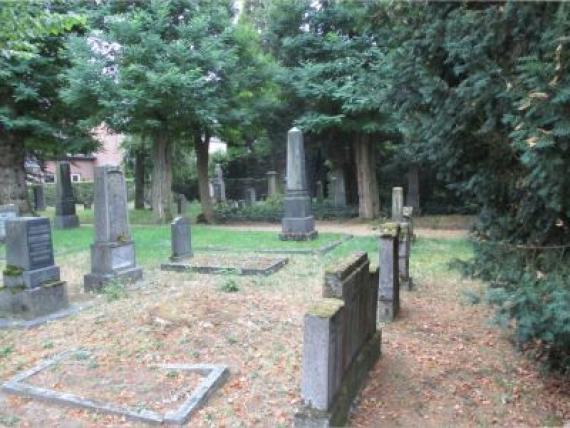 Farbfotografie von einigen Gräbern auf dem Friedhof. Rechts und im Hintergrund sind Bäume und Büsche zu erkennen.