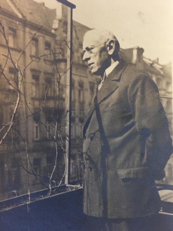 Homeowner Isidor Lewy, in a suit and glasses, stands at the balcony parapet of his house with his arms folded behind his back, looking through a flower grate at the opposite side of the street.