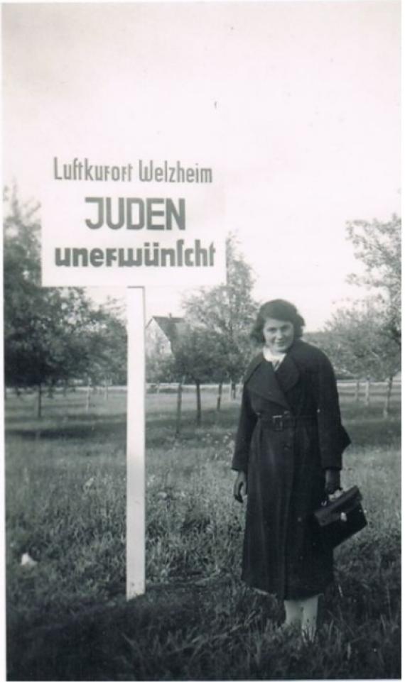 Eine junge Frau (Helene Adler) steht vor ein großen weißen Schild. Ganz oben auf den Schild  steht: "Luftkurort Welzheim"
Drunter steht: "Juden unerwünscht". 