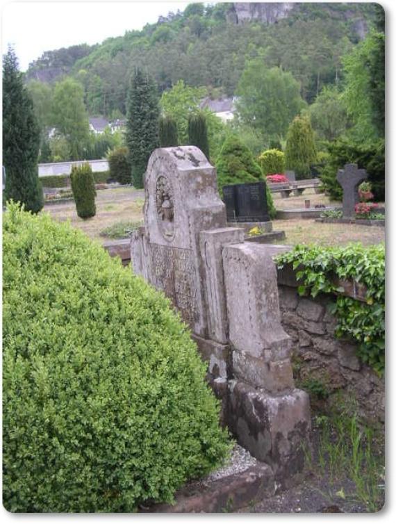 High gravestones stand behind a hedge. Mountains are visible in the background.