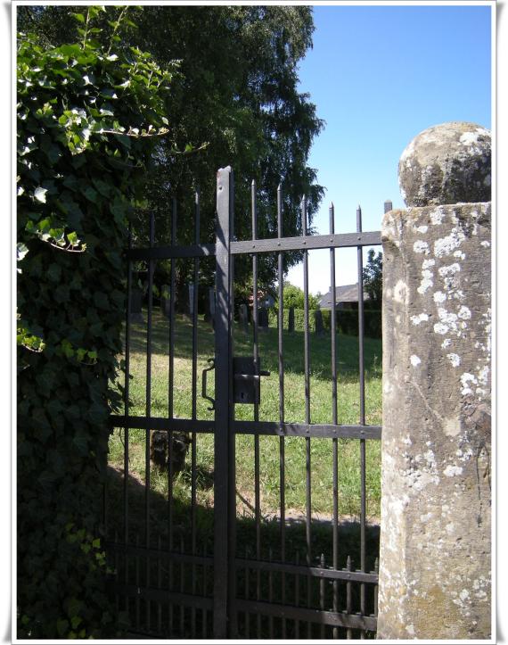 Friedhofseingang durch ein metallenes Gitter. Rechts ist ein Gemäuer zu sehen.