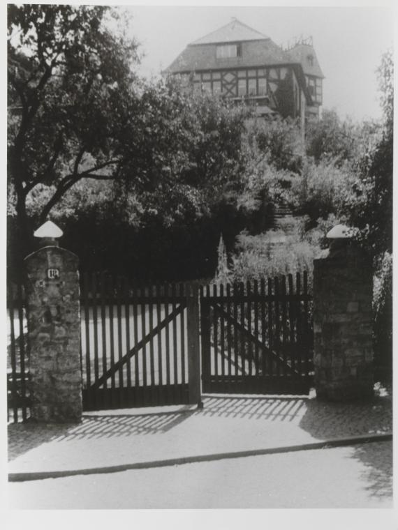 You can see a slightly open gate that leads to the home. In the upper half of the picture you can see the roof of the home, but the rest of the house is hidden by trees.