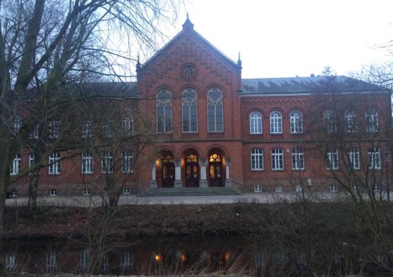  Current photo of the Old Grammar School. You can see a building made of red-brown brick. There are four columns in front of the three entrance doors, which can be reached through an open staircase. In front of the building there is a river surrounded by some trees.