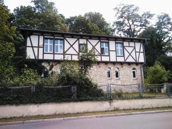 A half-timbered house photographed from the side from the street. You can see several modern windows, but some windows were left in the older style. The house is surrounded by trees from the left side.