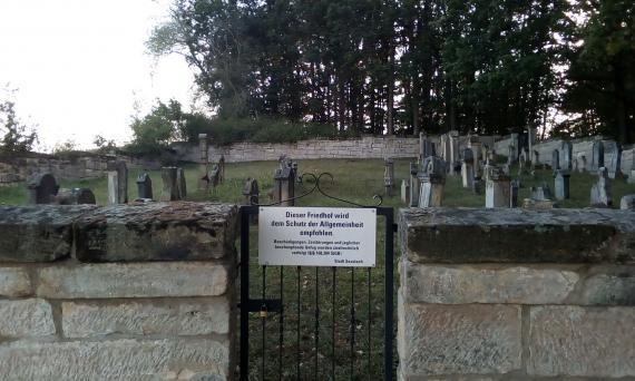 The photo shows the entrance gate of the Jewish cemetery in Autenhausen (Seßlach). On the right and on the left the cemetery wall goes off. In the background some gravestones can be seen on a lawn.