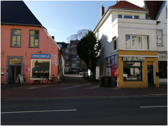 Auf dem Bild sieht man die Auffahrt zur Hausnummer 30, die in zweiter Reihe zur Straße steht. Neben der Auffahrt sieht man zwei Geschäfte.