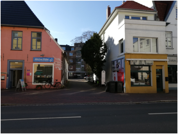 Auf dem Bild sieht man die Auffahrt zur Hausnummer 30, die in zweiter Reihe zur Straße steht. Neben der Auffahrt sieht man zwei Geschäfte. 