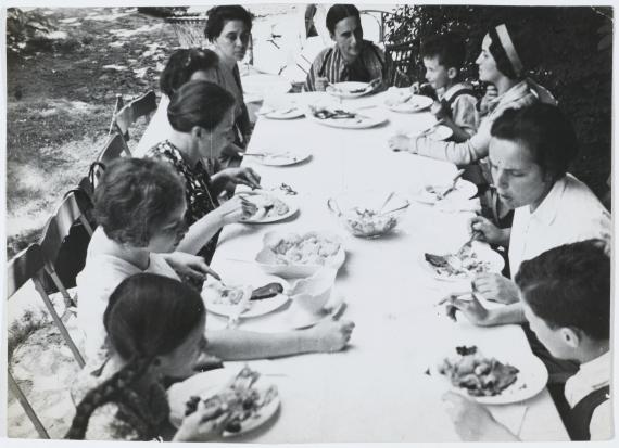 Schwarz-Weiß-Foto mit einer langen Tafel im Garten an der Frauen und Kinder zum gemeinsamen Essen sitzen und sich rege unterhalten
