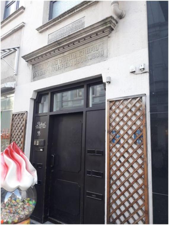 You can see a white painted residential house with a dark door. Above are two inscriptions embedded in the wall of the house. They give information about the construction and the architects.