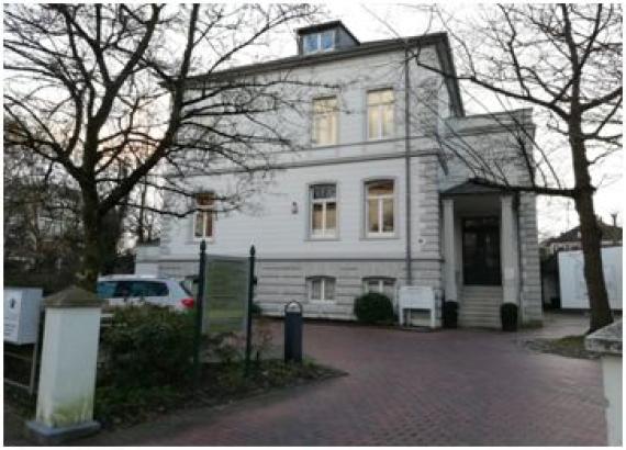 Color photo of the former home of the Reyersbach family. You can see a three-story white brick building, which is set off below with light gray stones On the right is the roofed house entrance.