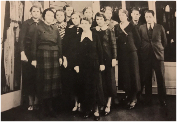 Das Bild zeigt Wallheimers Angestellte (11 Frauen und 2 Männer). Im Hintergrund sind eine Fensterfront und ein Kleiderregal zu sehen.