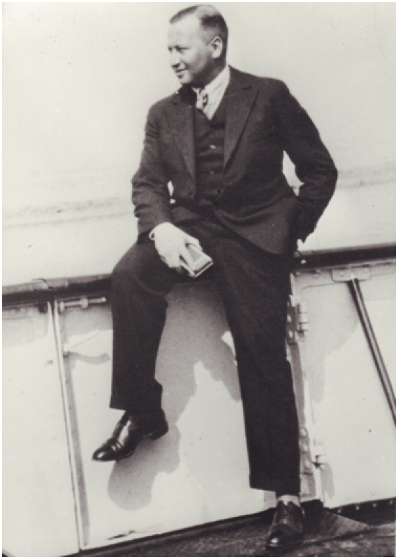 Bruno Wallheimer in a suit. He sits half on a railing, with one leg he supports himself on the ground