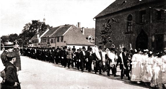 Schwarz-weiß Fotografie des Festzuges zur Einweihung der neuen Synagoge 1906. An der Zugspitze laufen einige junge Frauen in hellen, langen Gewändern. Im Anschluss folgen zahlreiche Männer mit Frack und Zylinder.