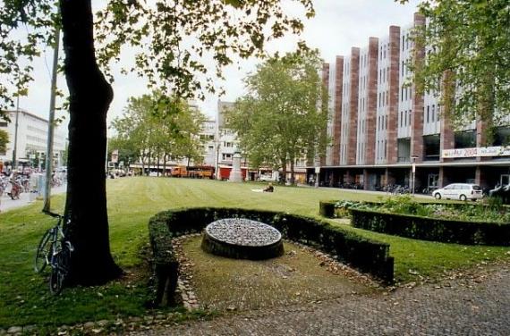 Der mit Gras und Bäumen bewachsene Platz der Alten Synagoge vor dem Umbau 2016. Im Vordergrund ist die Gedenktafel für die Zerstörung der Synagoge zu sehen.