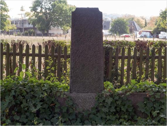 Ein einzelner verbliebener Grabstein ohne Innschrift. Der Stein steh vor einem hüfthohen Holzzaun, hinter dem sich ein Feld erstreckt.