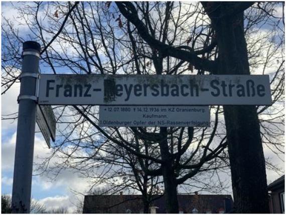 Leicht vermoostes Straßenschild in Gedenken an Franz-Reyersbach in Oldenburg-Kreyenbrück. Unter dem Straßennamen ist ein kleines Schild mit Informationen zum Geburts- und Sterbedatum sowie Beruf (Kaufmann) und der Zusatz: „1. Oldenburger Opfer der NS-Rassenverfolgung“ zu sehen.