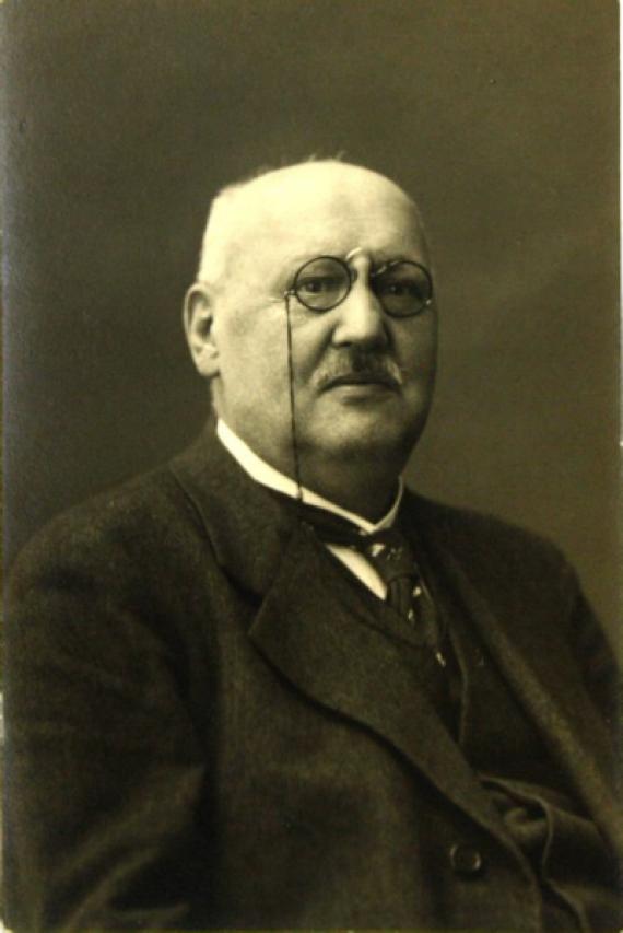 Black and white photograph by Adolf Bandmann. You can see an older gentleman with a strong build and little gray-white hair who wears a pince-nez and a suit and also has a gray mustache.