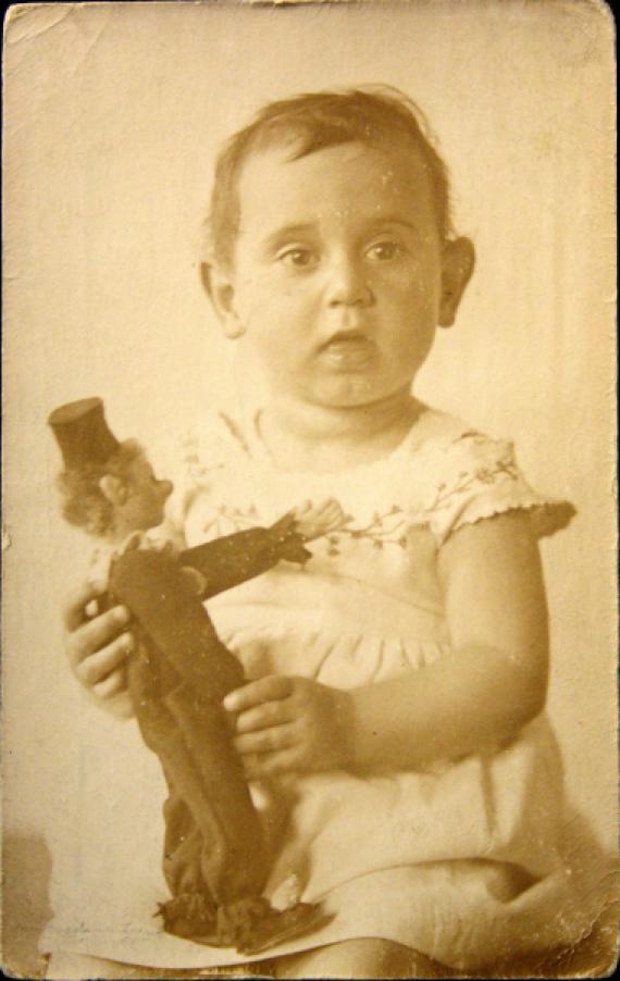 The toddler portrait shows Felice Schragenheim in a bright little dress with flower embroidery. She looks frontally, with open mouth, into the camera and holds a clown figurine in her hand.