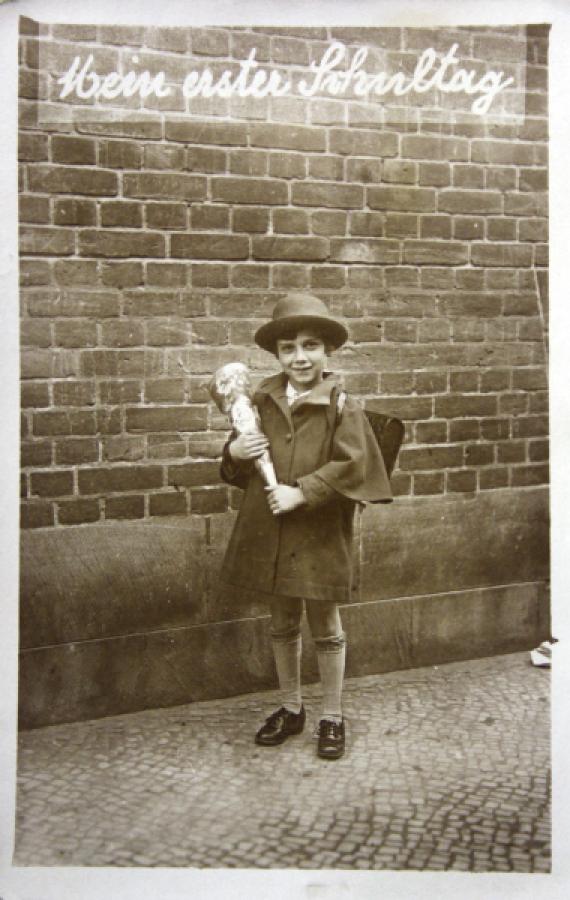 Felice Schragenheim is standing in front of a brick wall with a school bag and a satchel. The child is wearing a coat and a hat. The words "My first day at school" are inserted into the photograph at the top of the image.