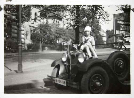 Felice Schragenheim sitzt auf der Kühlerhaube eines Autos. Sie trägt ein Kleid und einen Hut. Im Hintergrund ist ein Straßenzug mit Häuserfassaden und Bäumen zu sehen. Am rechten Bildrand ist eine Werbetafel zu erkennen. Die Aufnahme ist unscharf.