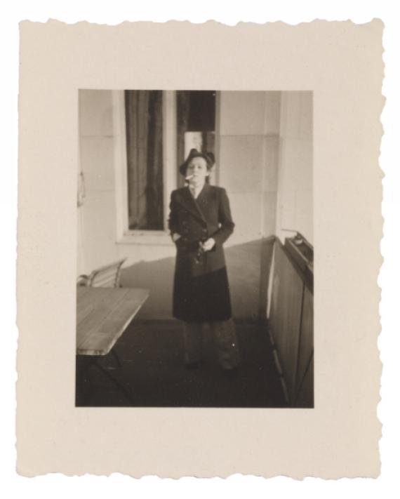 Felice Schragenheim is standing on a balcony with a cigarette in the right corner of her mouth. She wears a dark coat and a hat with wide pants. In the foreground a table can be seen, to the right the picture is bordered by a wall.