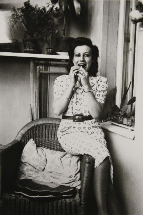 Felice Schragenheim is sitting on the back of a wicker chair on the balcony. With her hands she holds something to eat in front of her open mouth. She is wearing a light printed dress with a belt at the waist. To the right of Felice Schragenheim there is a window, in the background there are flower pots on a board on the wall.