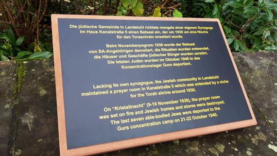 It is a black plaque with golden writing. The text is written in German and English.