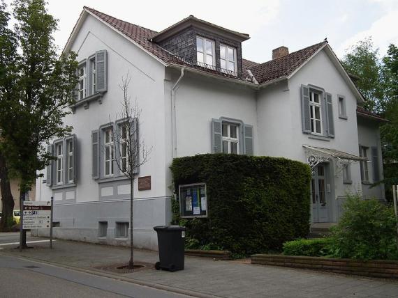 Außenansicht des Hauses mit einer hellen Fassade und geöffneten Fensterläden. Im rechten Teil des Bildes ist die Eingangstür hinter einer Hecke zu sehen. 