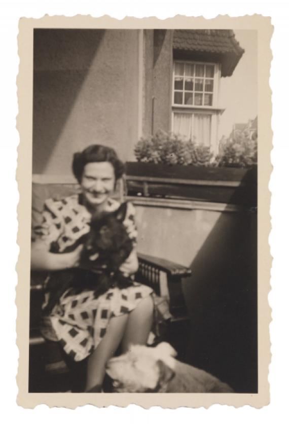 Irene wears a patterned dress and looks into the camera with a smile. In the background, a flower box on the balcony railing and the corner of a house can be seen. The shot is a bit out of focus.