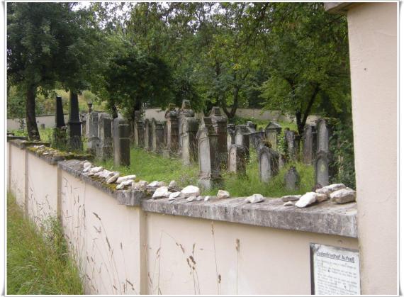 In the foreground a bright cemetery wall, behind it a lawn with gravestones under trees, gloomy mood.