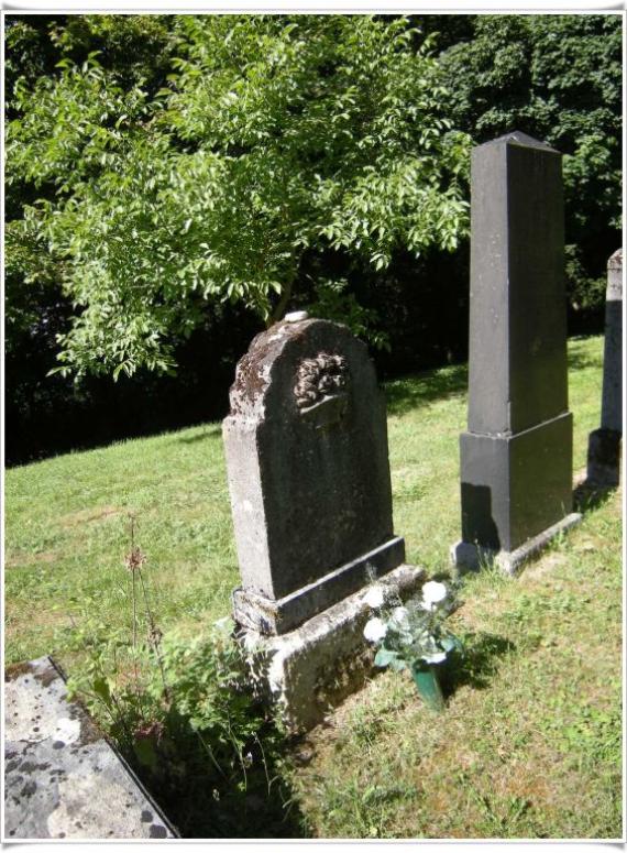 Two fully captured tombstones. One traditional with round arch, the other in obelisk form. Stones on the meadow, in the background forest.