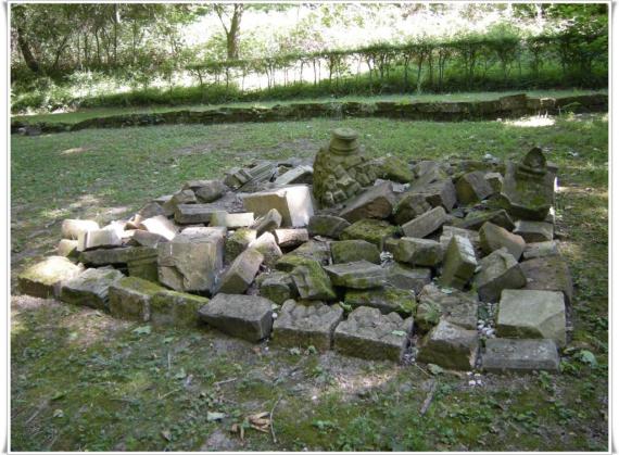 Wiese. Auf der Wiese viele Trümmer von Grabsteinen. Aufgeschichtet. Bilden ein Mahnmal.