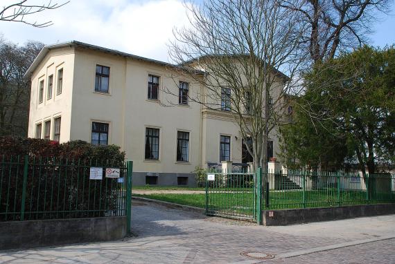 Frankfurt (Oder), Gubener Straße 15 A Vorderseite der gelblich geputzten zweistöckigen Villa mit Garten, Nebengebäude und Zaun. Man sieht ein Villengebäude mit zwei Seitenflügeln, Eingangstreppe am mittleren Gebäude. Rechts davon ein Nebengebäude, etwas Garten vor den Gebäuden und Zaun zur Straße.