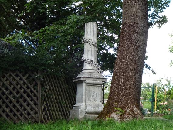 Grabstein mit Säule, rechts daneben ein Baum, links daneben ein Holzzaun