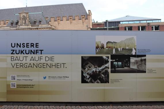 Bauzaun mit Plakatwand. Entwürfe des neuen Museums sind abgebildet.