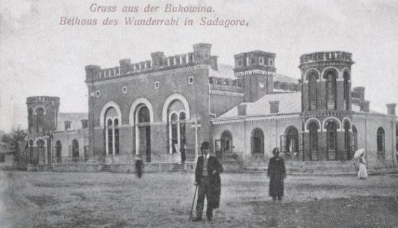 Old photo with a man in the foreground. He is wearing a suit and top hat and holding a cane. In the background is a large synagogue.