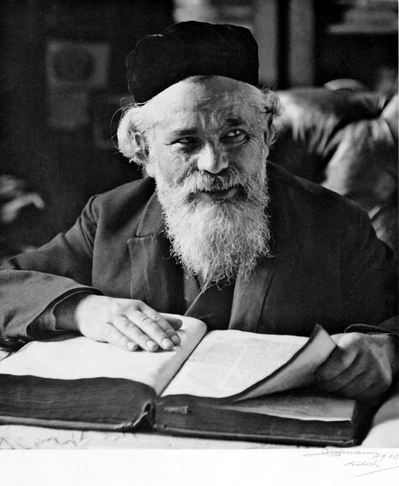 Old portrait of an elderly man sitting at a table. In front of him is a book and he has a beard.