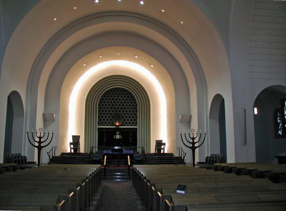 Interior of the synagogue. A shrine in the center, two menorahs on the sides and pews.