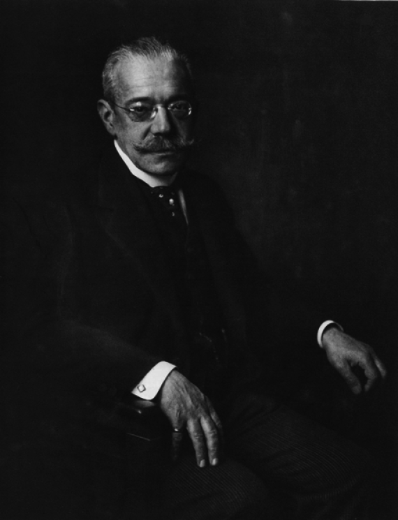 Old portrait of James Simon. He wears a suit and glasses.the shot is made from the side sitting.