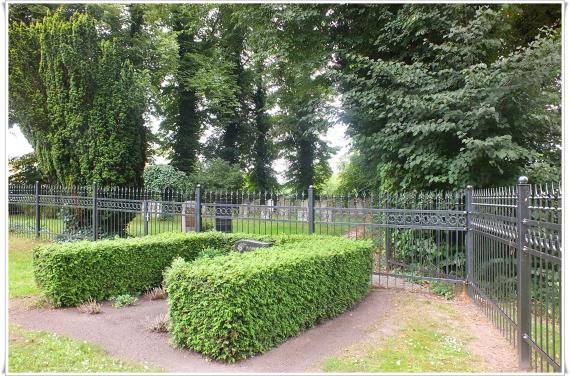 Ein Grab. Von einer halbhohen grünen Hecke umgeben. Im Hintergrund ein Zaun. Dahinter mehrere Grabsteine.