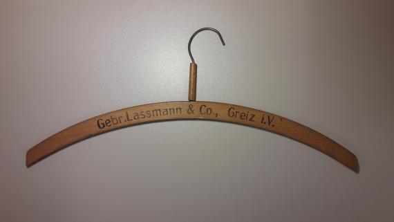 Man sieht einen hölzernen Kleiderbügel des Geschäfts "Gebrüder Laßmann & Co." mit einem Haken aus Metall. Der Bügel trägt die Beschriftung "Gebr. Lassmann & Co., Greiz i. V." (= im Vogtland).