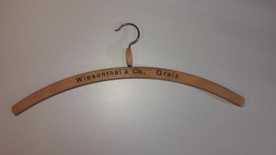 Man sieht einen hölzernen Kleiderbügel des Geschäfts "Wiesenthal & Co." mit einem Haken aus Metall. Der Bügel trägt die Beschriftung "Wiesenthal & Co., Greiz".