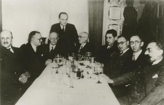 Nine men grouped around inn table with wine glasses