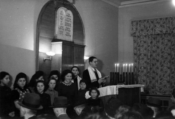 Service in a small synagogue. Six Hanukkah candles burn on the prayer lectern, the cantor prays, ten young people stand opposite the congregation