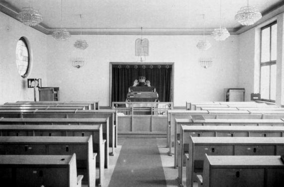 Simple prayer room, central aisle between rows of benches, background lectern and velvet curtain in front of the wall niche with the Aron Hakodesh.