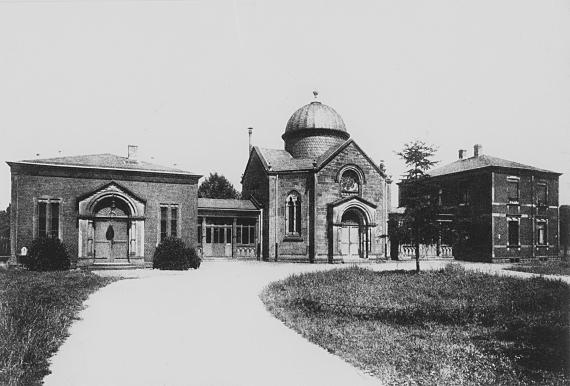 Three buildings, connected with corridors, the middle one with dome. In the foreground access road with green area