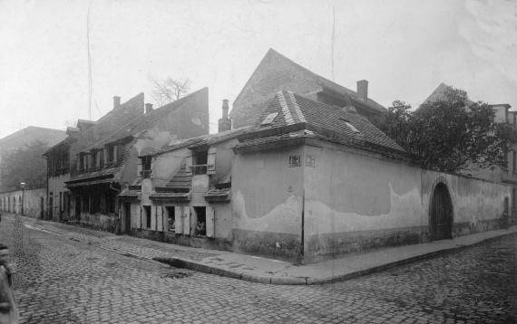 Straßenecke mit kleinen Häuschen, dahinter Bäume, die aus dem alten jüdischen Friedhof hervorragen