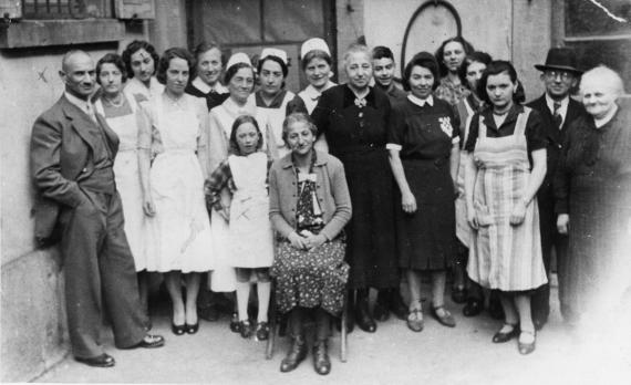 Personengruppe verschiedener Altersstufen in einem Hinterhof. Einige jüngere Frauen tragen Krankenschwesternkleidung.